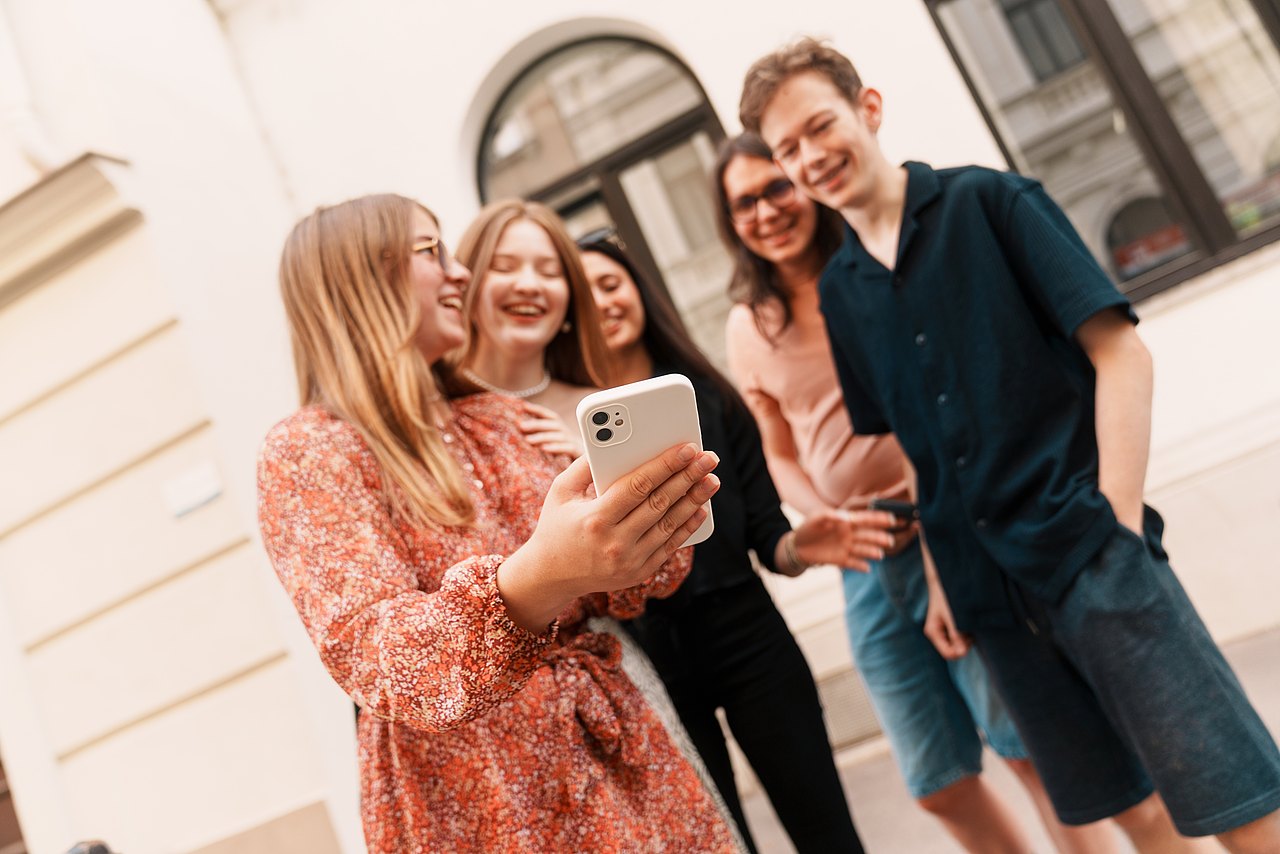 mz2025 x ste.krenn-216 Eine Gruppe an lachenden Jugendlichen steht vor einem Haus in einem Kreis und schaut auf ein Handy..