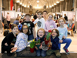 Eine fröhliche Kinderschar freut sich und einige Kinder halten Blumentöpfe oder Tischtennisschläger in den Händen.