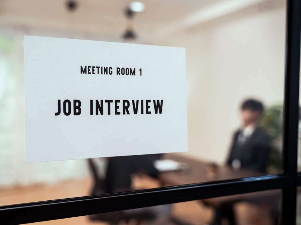 Auf einer Glastür hängt ein Schild mit "Job Interview" dahinter sieht man verschwommen ein Bewerbungsgespräch