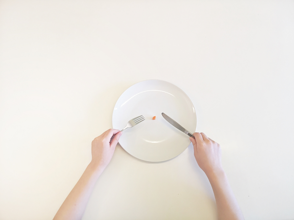 Ein Teller mit einer Person die Besteck in der Hand hält - auf dem Teller liegt ein sehr kleines Stück essen