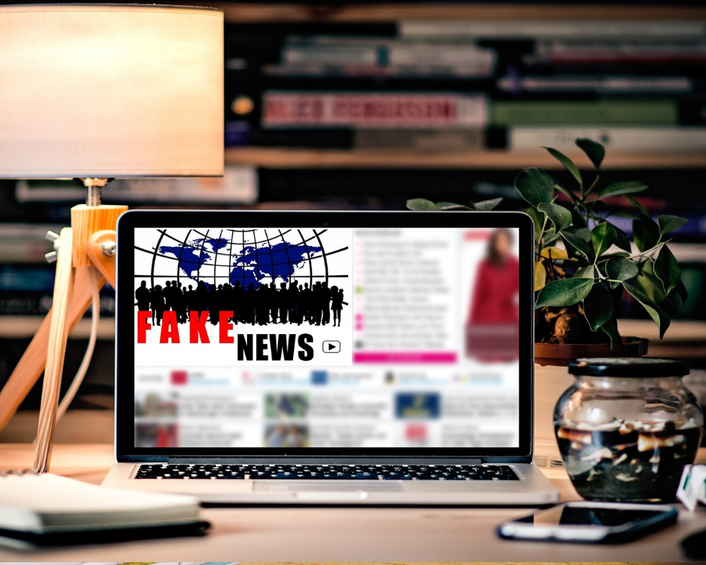 Ein Laptop auf einem Schreibtisch mit Bildschirmhintergrund zu "Fake News", daneben steht eine Pflanze und dahinter ist ein Bücherregal