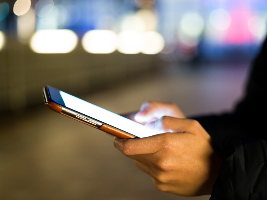 eine Person hält ein Handy in der Hand. Im Hintergrund sieht man die Nachtlichter einer Stadt
