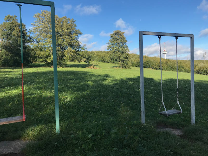 Das Bild zeigt zwei einfache Schaukeln auf einer Wiese im Freien. Die Schaukeln hängen an Metallgestellen und bestehen aus schlichten Sitzbrettern. Sie stehen nebeneinander auf einer grünen Rasenfläche. Im Hintergrund sieht man Bäume, Sträucher und eine leicht hügelige Landschaft unter einem blauen Himmel. Die Umgebung wirkt ruhig, sonnig und naturnah – ein offener Platz, der viel Raum zum Spielen und Schaukeln bietet.