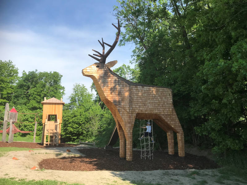 Hirsch aus Holz Riesen-Holzhirsch mit Rutsche. Im Hintergrund ein Kletterturm.