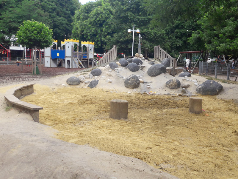 Ein Hügel aus Beton mit mehreren kleineren und großen Steinen. Ganz oben ist eine Wasserpumpe und es führt ein Weg für das Wasser hinab. Daneben ein Sandspielbereich mit einer Holzbank und zwei Holzstämmen.