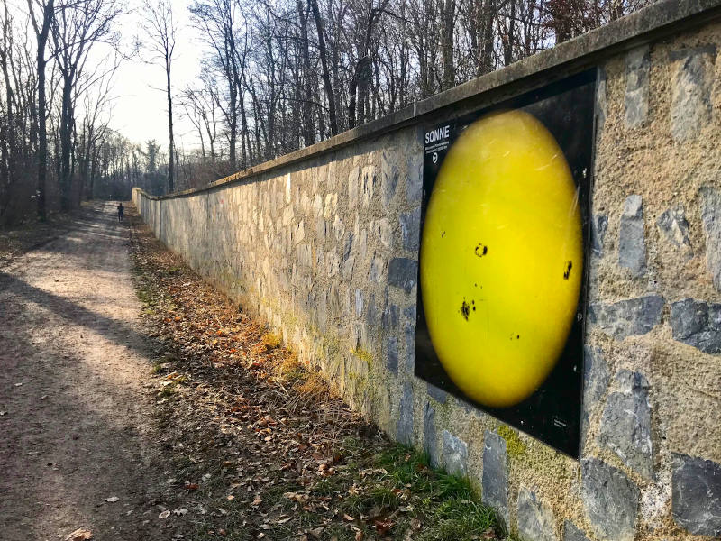 Pfad in der Natur, daneben eine Steinmauer mit einer Abbildung der Sonne.