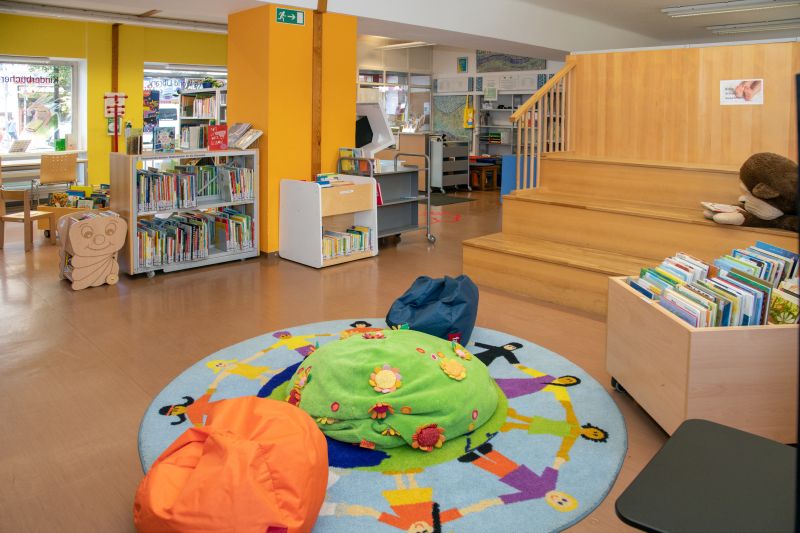 Teppich Bunter Teppich mit Sitzsäcken und einer Bücherkiste im Vordergrund, dahinter Holzstiegen und mehrere Bücherregale.