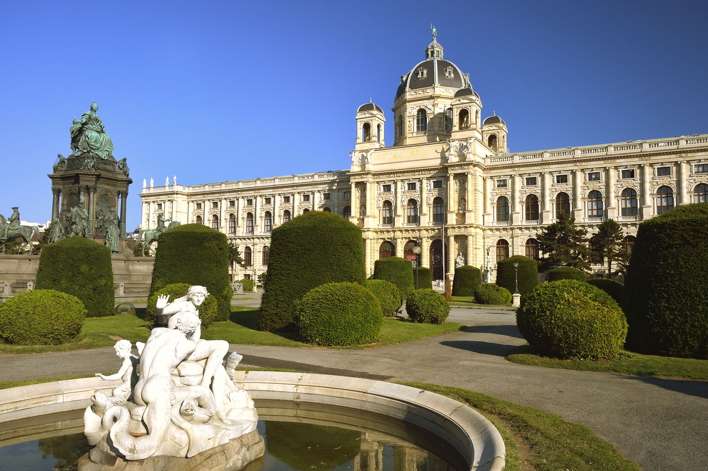NHM Im Vordergrund ein Brunnen mit Statue, im Hintergrund das Naturhistorische Museum von vorne, dazwischen die Wege und Hecken des Maria-Theresien-Parks. Ganz links die Statue von Maria Theresia.