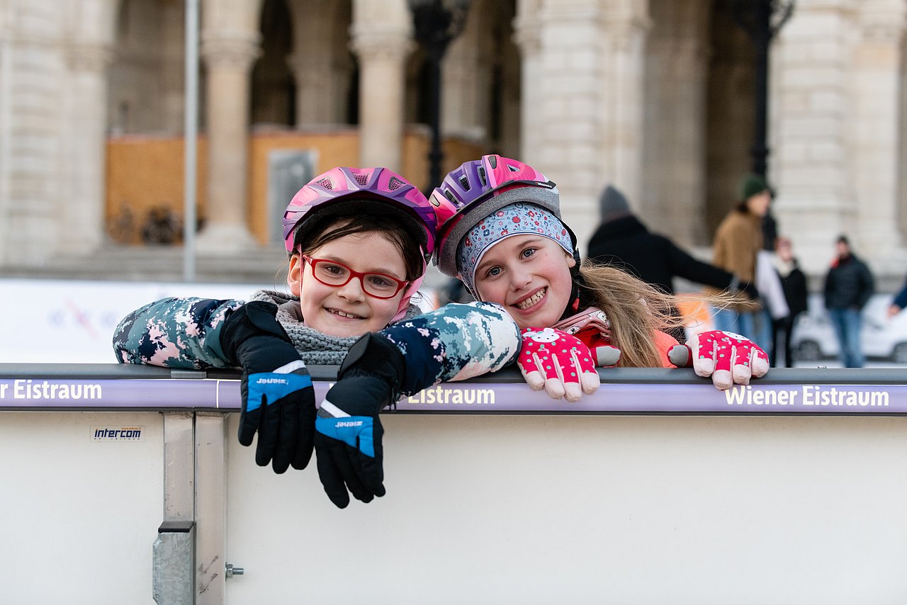 Wiener Eistraum Zwei Mädchen die in Eislaufausrüstung bei der Wand des Eislaufplatzes lehnen