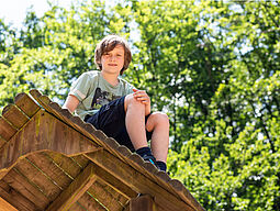 Abenteuer_Wald_4 Bub sitzt auf dem Dach einer Holzhuette.