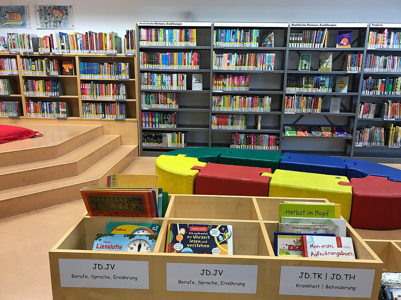 Bücherei Gegenüber eine Bücherwand, links eine Holzstiege, in der Mitte vom Raum bunte Puzzle-Sitzsteine die ein Oval bilden, davor Bücherkisten.