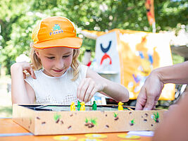 Zu sehen ist ein Kind auf einer Spieleveranstaltung im Park. Das Kind sitzt an einem Spieltisch. Gegenüber sitzt eine weitere Person, von der nur eine Hand zu sehen ist. Sie spiele das Spiel Maulwurf Company und das Kind macht gerade einen Spielzug.