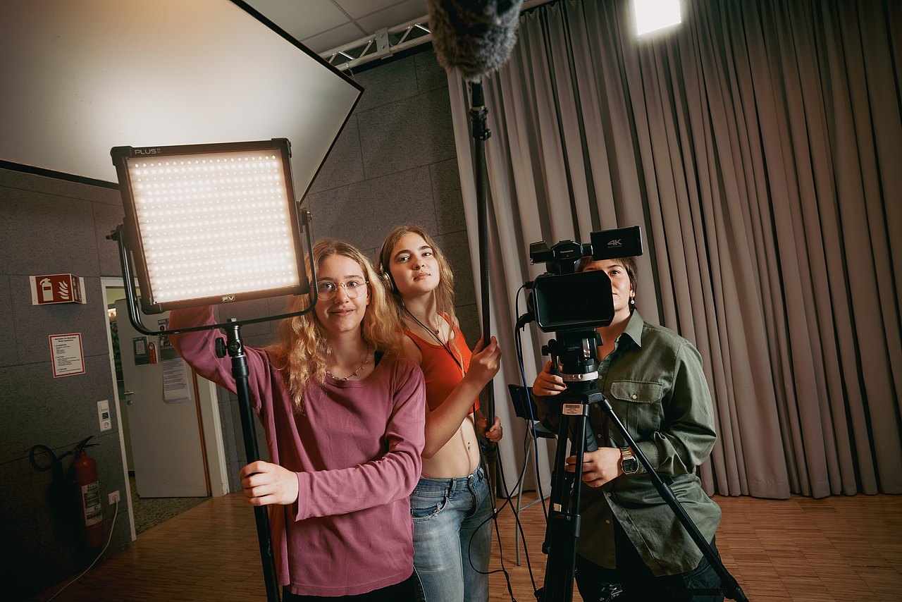 MZ_23_KRENN_v1-144 Eine Jugendliche bedient eine Panasonic-Videokamera in einem Studio, während eine andere Jugendliche eine LED-Flächenleuchte ausrichtet und eine dritte Jugendliche ein Mikrofon hält.