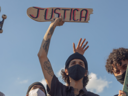 3 Menschen von unten fotografiert, Hintergrund blauer Himmel. Eine Person hält zerbrochenes Skateboard mit Aufschrift "Justica"