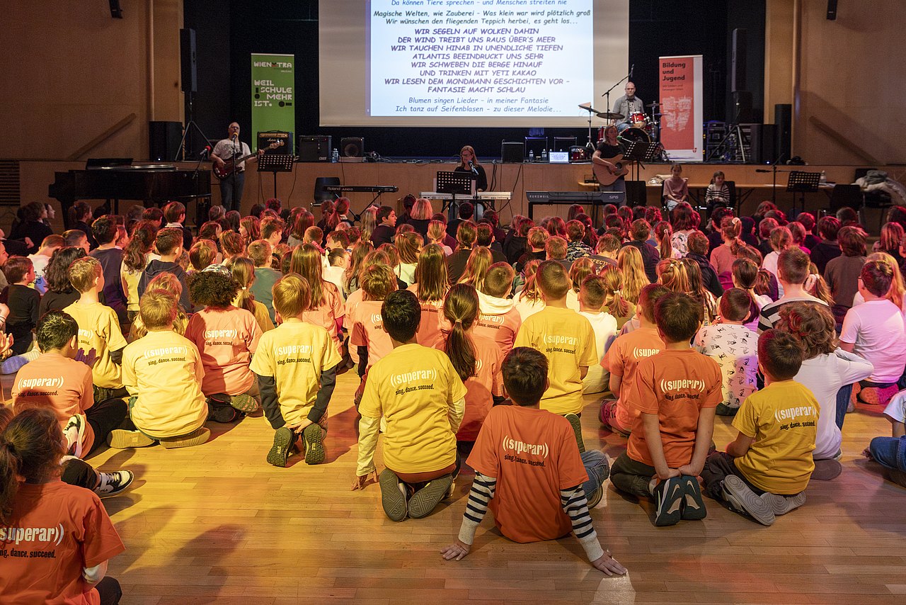 WienXtra, Musik aktiv-c-Carolina Frank-104 Große Gruppe Kinder sitzt am Boden und singt gemeinsam mit Band