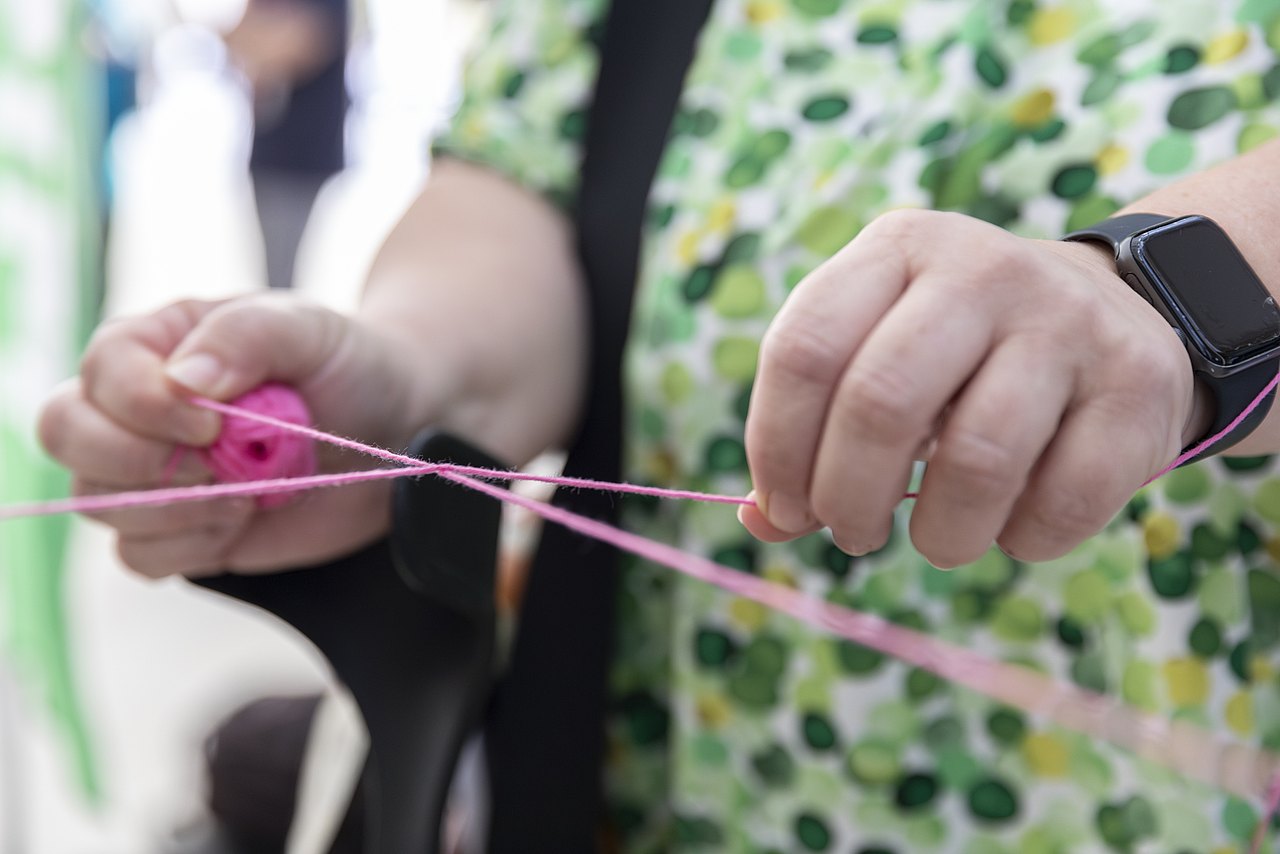 20230920_IFP Tagung-c-Carolina Frank_05_Gegenstand_kein Gesicht Die rechte und linke Hand einer weißen Person ziehen an zwei Enden von einem rosa Faden und bilden damit einen Konten.