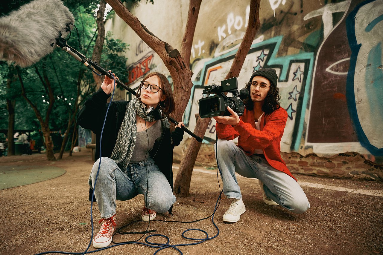 MZ_23_KRENN_v1-38 Zwei Jugendliche knien in einem Park am Boden, einer der Jugendlichen hält eine Videokamera, während die andere Jugendliche ein Mikrofon hält.