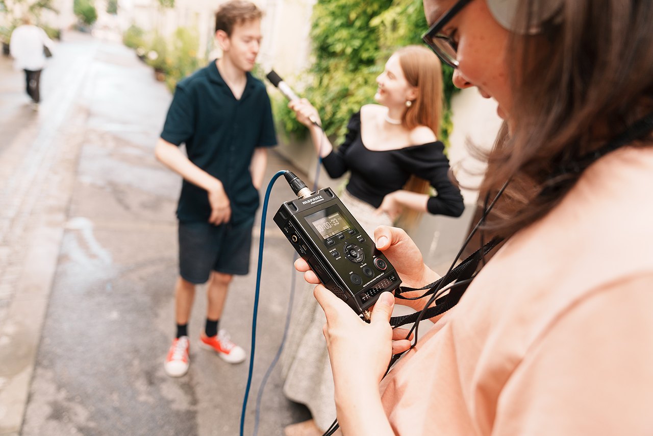 mz2025 x ste.krenn-208 Ein Jugendlicher hält ein tragbares Audiorekorder-Gerät, während zwei andere Jugendliche ein Interview mit einem Mikrofon aufnehmen.