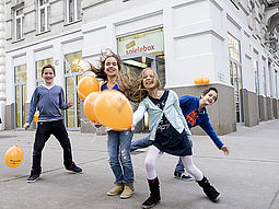 Kinder mit Luftballons springen vor der wienXtra-spielebox herum