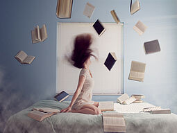 Junge Frau sitzt im Bett, rund um sie herum fliegen Bücher durch die Luft