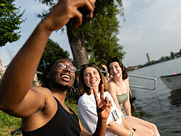 Jugend Drei Jugendliche sitzen am Wasser und machen ein Selfie.