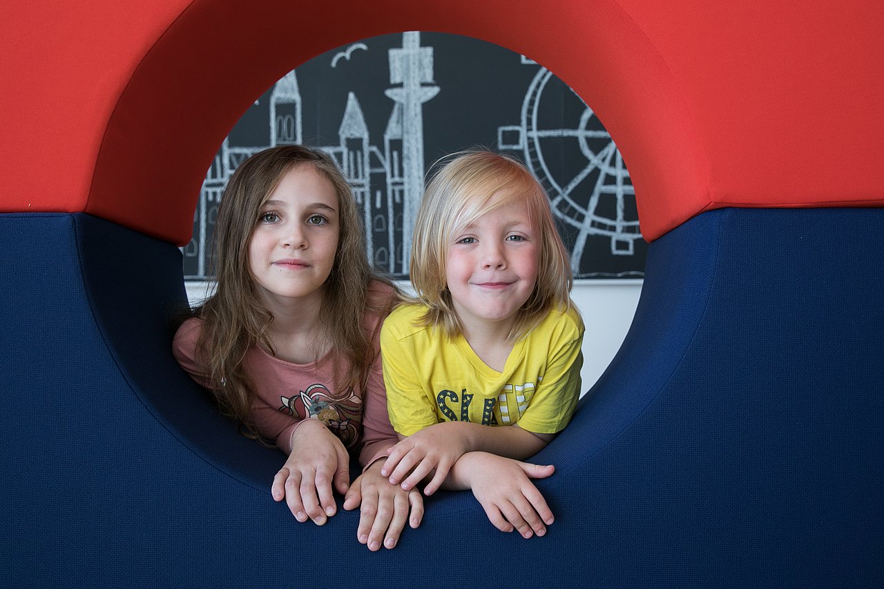 Kinder, die durch ein rundes Fenster schauen und lächeln, im Hintergrund eine Zeichnung vom Donauturm