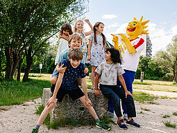 Sommer Ferienspiel-Maskottchen Holli lacht gemeinsam mit 6 Kindern.