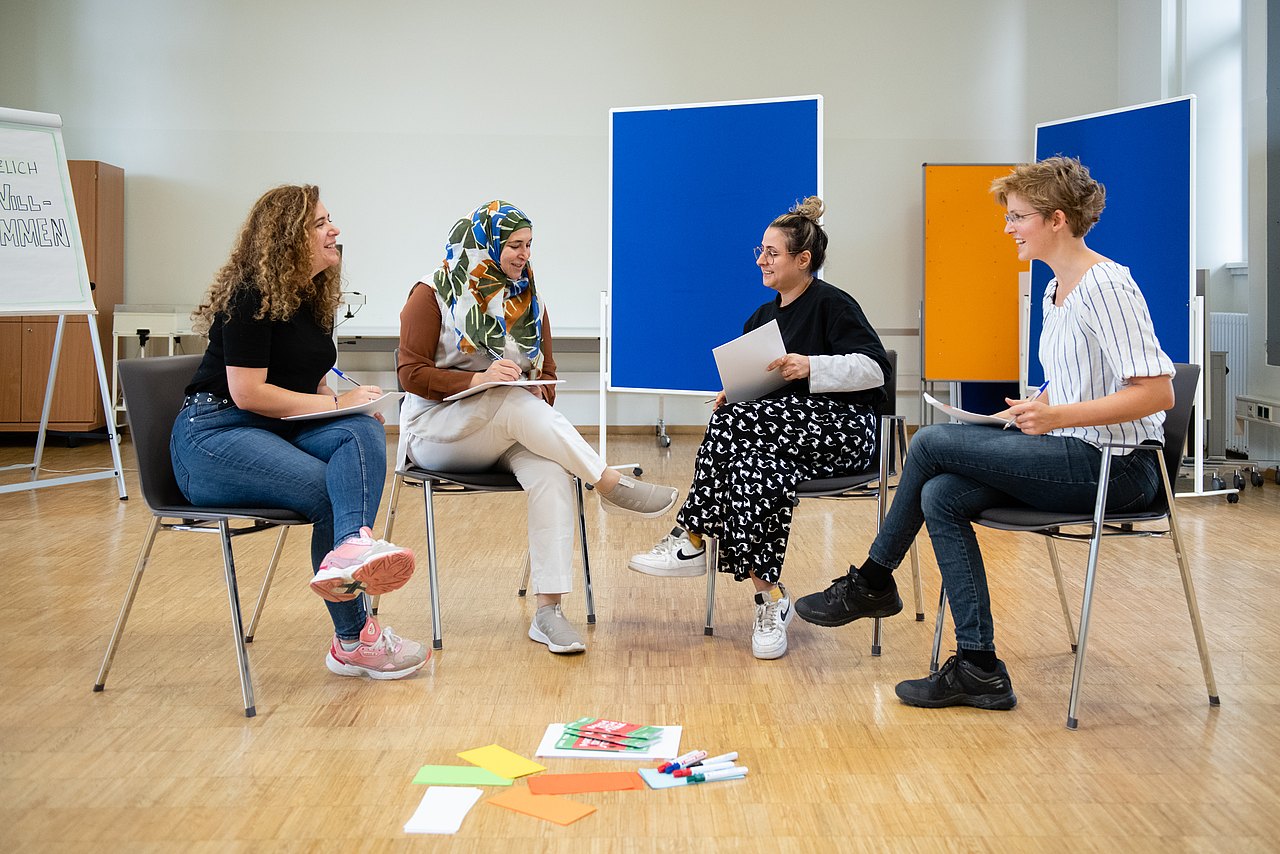 Praxisnahe Weiterbildung, die begeistert! Vier Frauen sitzen auf Sesseln im Kreis im Seminarraum und sprechen miteinander. Am Boden liegen Moderationskärtchen und im Hintergrund sind verschiedenfarbige Pinnwände aufgestellt