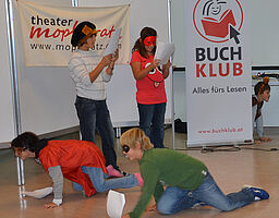 Zwei Kinder lesen stehend einen Text, zwei Kinder krabbeln am Boden, alle tragen Masken
