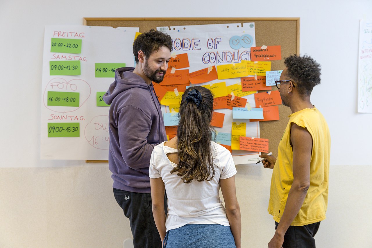 Hier entstehen Ideen Drei Personen unterhalten sich vor einer Pinnwand, auf der Post-its hängen