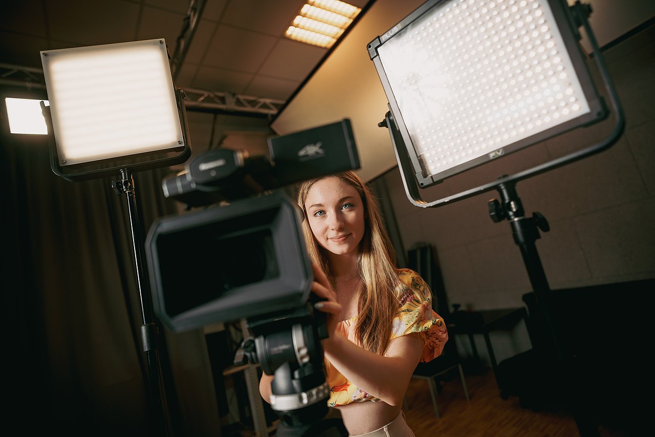 MZ_23_KRENN_v1-146 Eine Jugendliche steht hinter einer Kamera auf einem Stativ und zwei Videoleuchten in einem Raum.