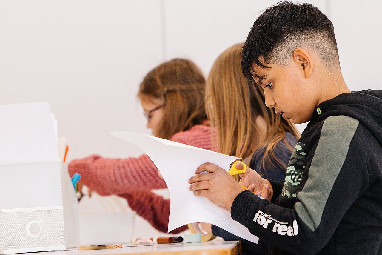 20250409-_MG_1173-©AlekKawka Kind schneidet Blatt Papier, dahinter zwei Kinder am kreativ arbeiten