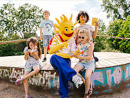 Sommer Ferienspiel-Maskottchen Holli blättert gemeinsam mit Kindern im Programm vom Ferienspiel.