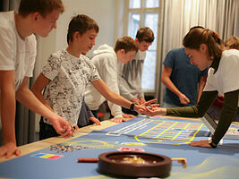 Schüler_innen stehen mit verschiedenem Material um Tisch herum, auf dem ein Spielplan liegt