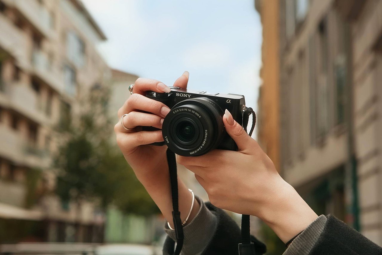 Eine Jugendliche hält eine kompakte Sony-Kamera in den Händen und fotografiert auf einer Straße zwischen Gebäuden.