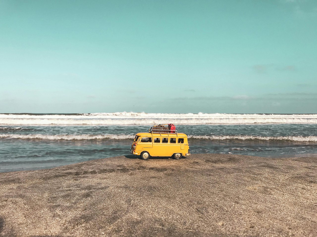 Gelber Spielzeug-Minibus mit Gepäck auf dem Dach steht am Strand vor dem Meer.