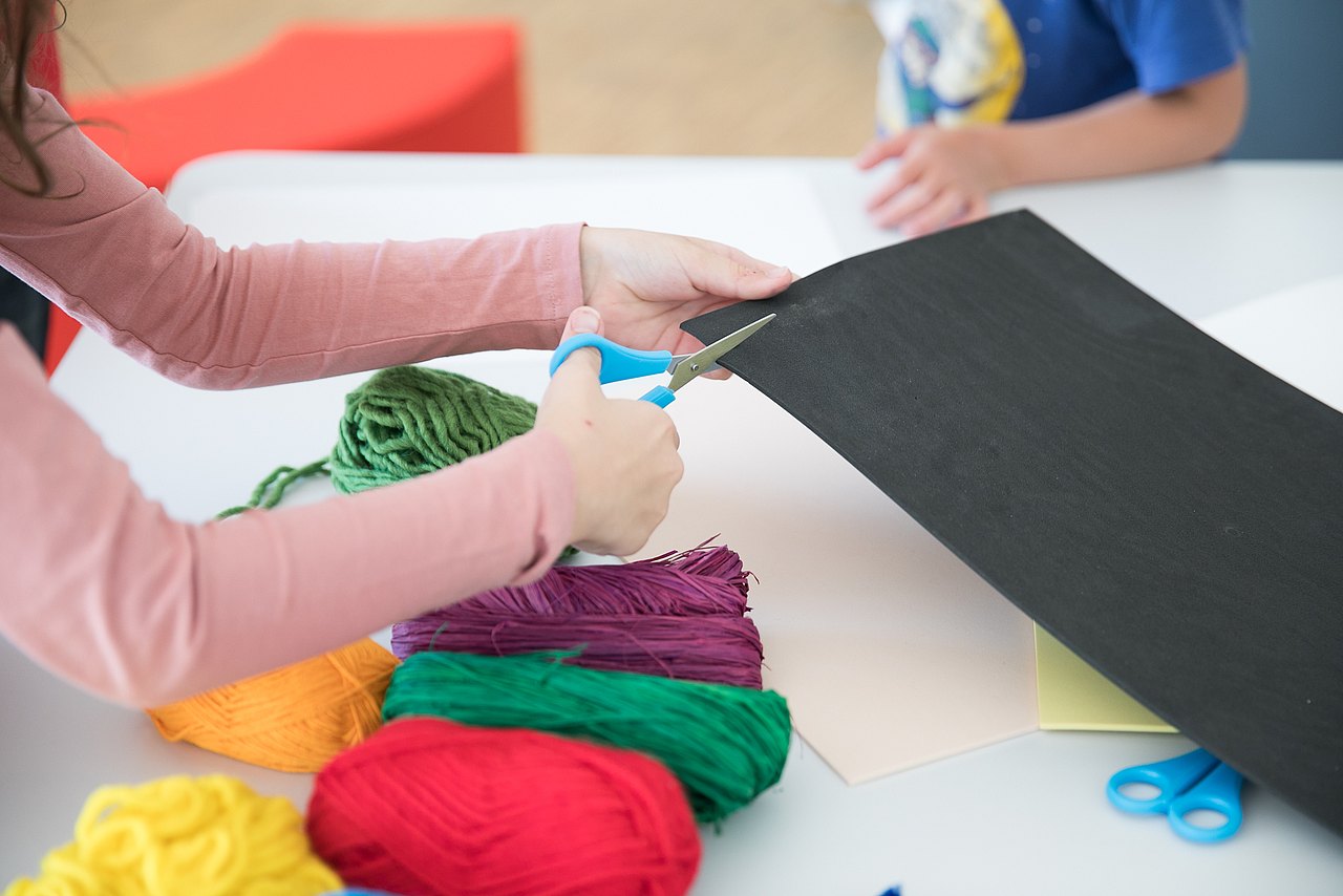 20210903_IMG_1874 Kind schneidet mit Schere Blatt Papier, Wollknäuel liegen auf dem Tisch