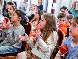 Drei Mädchen im Vordergrund und weitere Kinder in den hinteren Reihen applaudieren. 