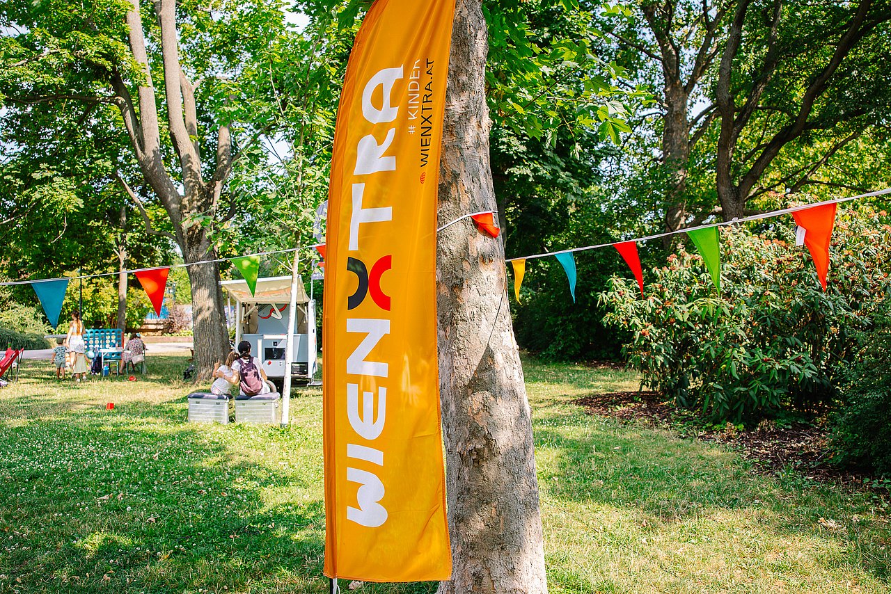 Hollis Grätzltour und Vorhang auf im Park orange Wienxtra Flagge vor Baum im Park