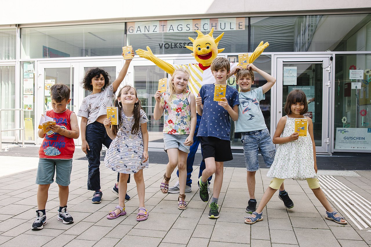 Kinderaktiv Ferienspiel fröhliche Kinder und Ferienspiel-Maskottchen Holli vor einer Schule