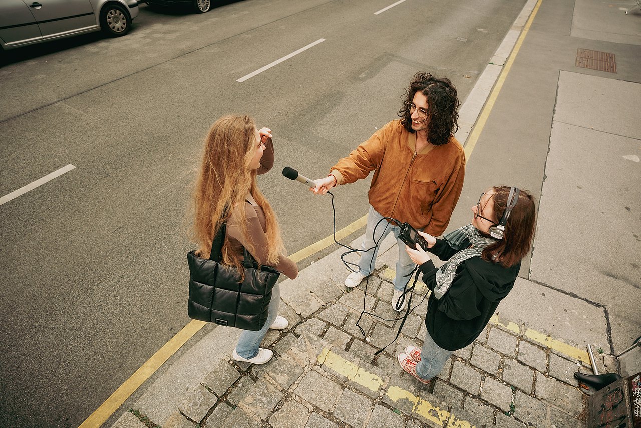 MZ_23_KRENN_v1-90 Drei Jugendliche stehen am Gehweg, einer hält ein Mikrofon und interviewt die andere Person.