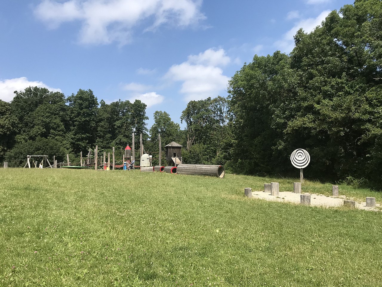Waldpädagogischer Spielplatz Am Himmel Spielgeräte auf Wiese mit daran angrenzendem Wald im Hintergrund