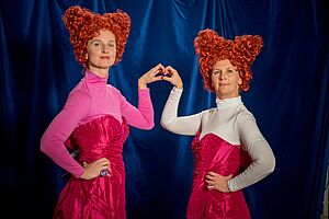 Zwei Schauspielerinnen in pinken schulterfreien Kleidern, mit rosa bzw. weißem Oberteil, und herz-förmigen Haarperücken, formen ihre Hände gemeinsam zu einem Herz
