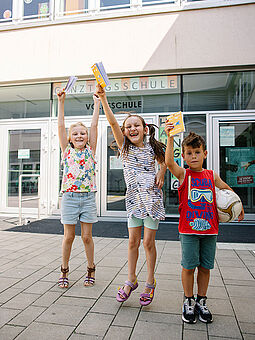 Sommer Zwei Mädchen und ein Bub hüpfen vor dem Eingang der Schule mit dem Ferienspielpass in der Hand.