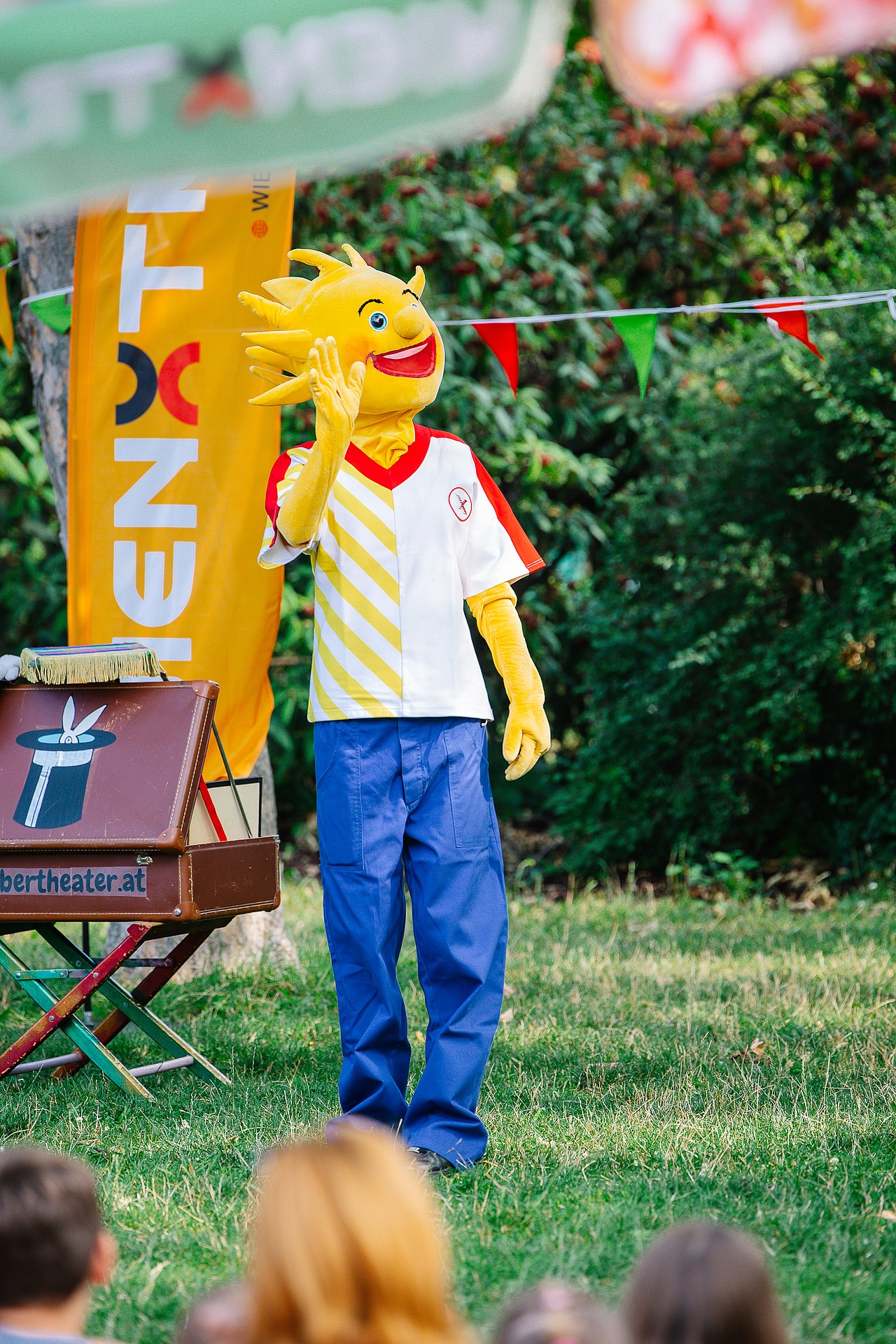 Hollis Grätzltour und Vorhang auf im Park Das Kinderaktiv-Maskottchen Holli steht im Park neben einem Zauberkoffer vor einer Wienxtra-Flagge und bunten Wimpeln und winkt