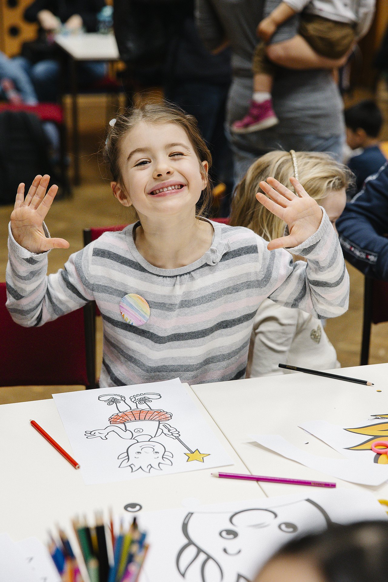 250228_MG_0825 breit lachendes Mädchen mit teils angemalten Malvorlagen an einem Tisch, beide Hände in die Luft gestreckt