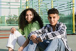 Ein Jugendlicher im karierten Hemd und Jeans und eine Jugendliche mit grünem Hemd und weißer Hose sitzen lässig am Boden vor einem Ballspielkäfig im freien. Sie blicken freundlich in die Kamera. 