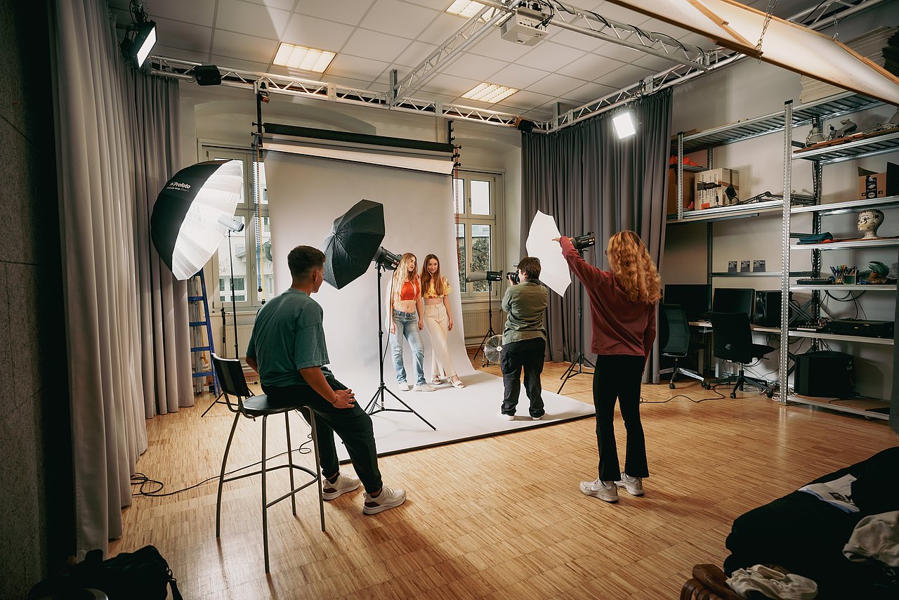 Zwei Jugendliche posieren in einem Fotostudio vor einem weißen Hintergrund, während eine dritte Jugendliche sie fotografiert, eine vierte ihnen Anweisungen gibt und ein fünfter Jugendlicher zusieht.