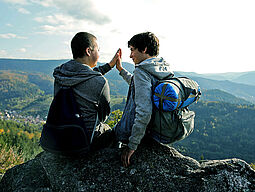 Zwei Freunde sitzen auf einem Berggipfel und geben sich ein High Five.