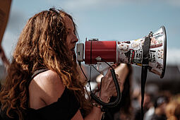 Person mit langen Haaren spricht mit Megaphone auf Kundgebung